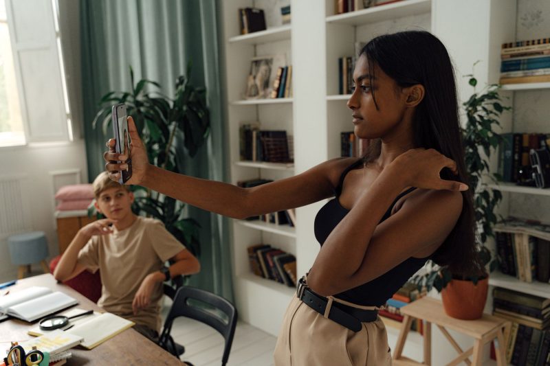 A woman takes a selfie in a stylish, plant-filled study while a young boy watches.