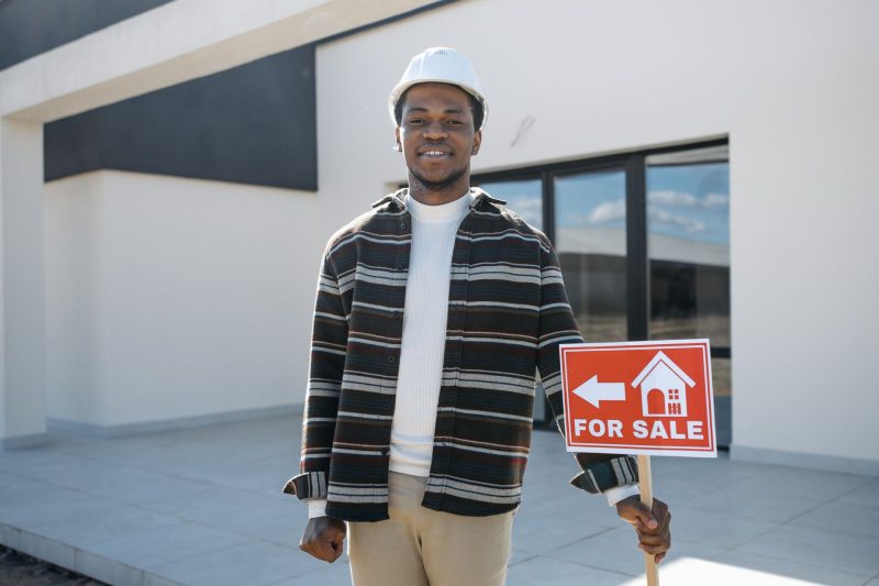 Home 18 Young black male realtor smiling while holding a "for sale" sign in front of a modern house.